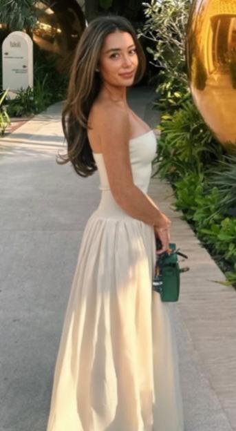 Jordan Porter, a management executive, standing on a paved path lined with lush green plants and large golden spherical objects, wearing a strapless white gown and holding a small green purse.