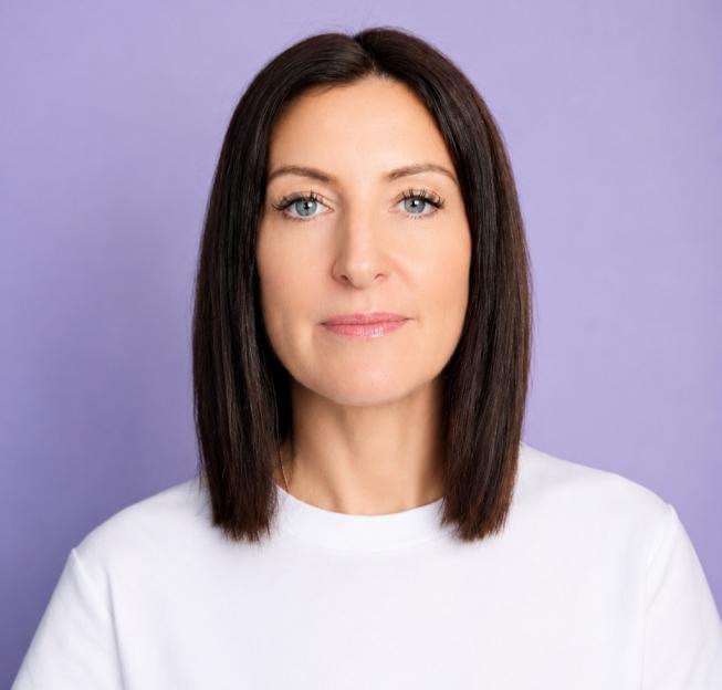A woman with dark hair and bright blue eyes, wearing a white t-shirt, stands against a lavender background.