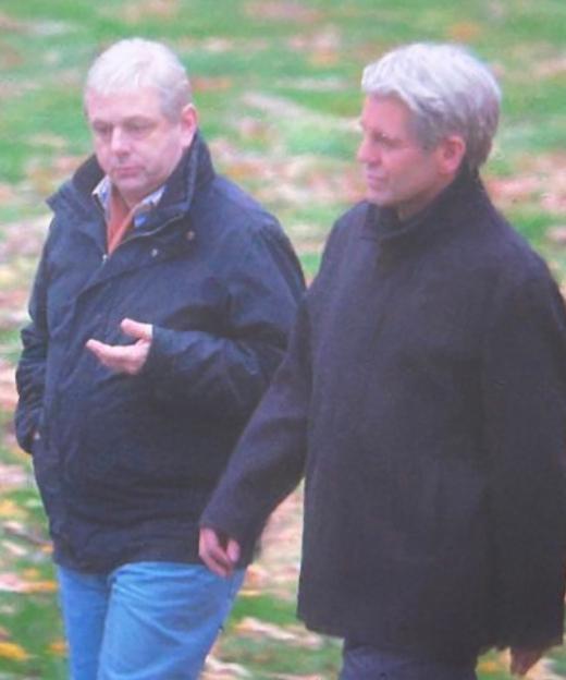 Two men walking outdoors, one wearing a navy coat and blue jeans, the other a black coat.