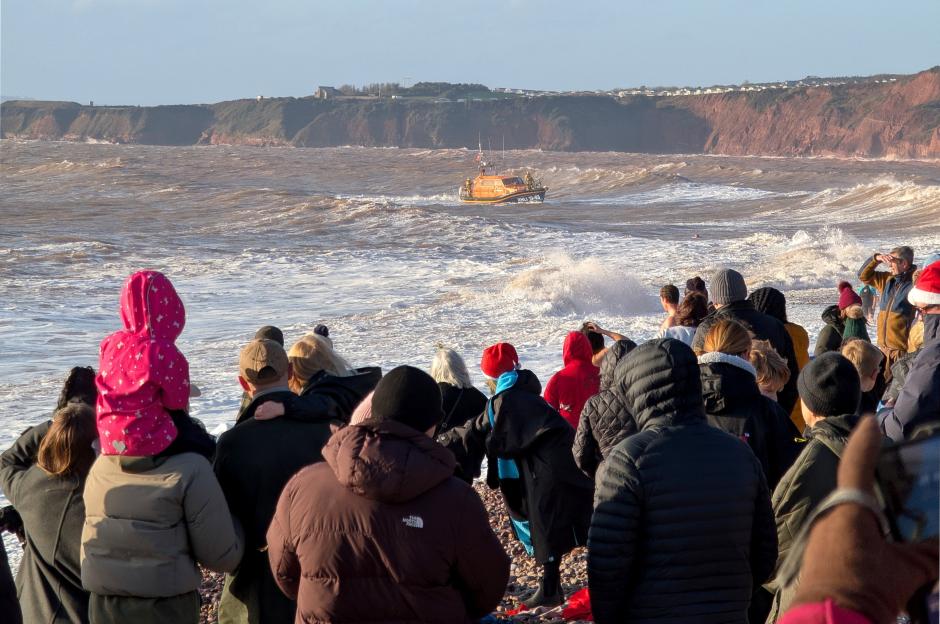Spectators on Budleigh Salterton beach watch a lifeboat in rough waters near cliffs.