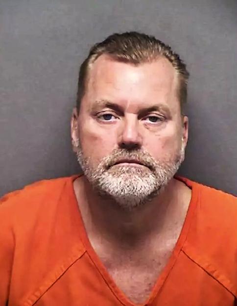 A man with light hair and a graying beard, wearing an orange jumpsuit, looks directly at the camera.