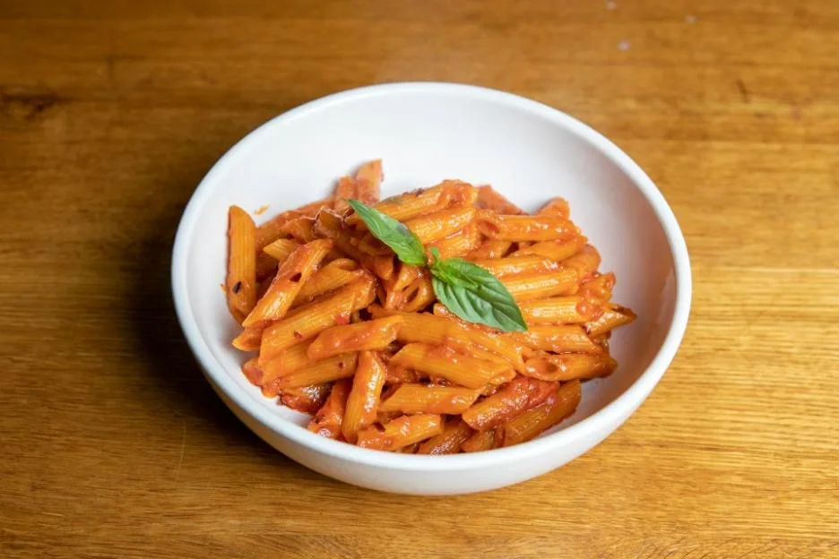 A bowl of penne arabiata garnished with basil leaves on a wooden table.