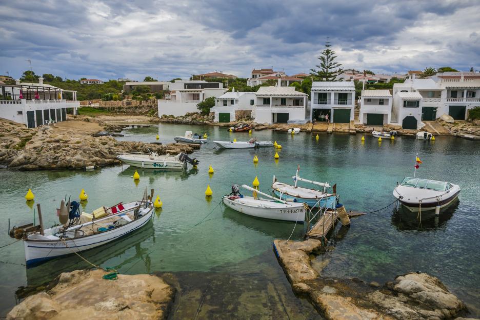 Cala Biniancolla beach with boats in the turquoise sea and white houses along the shore under a cloudy sky.