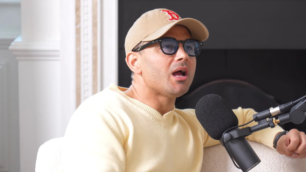 A man with a tan baseball cap, sunglasses, and a yellow sweater speaks into a microphone.