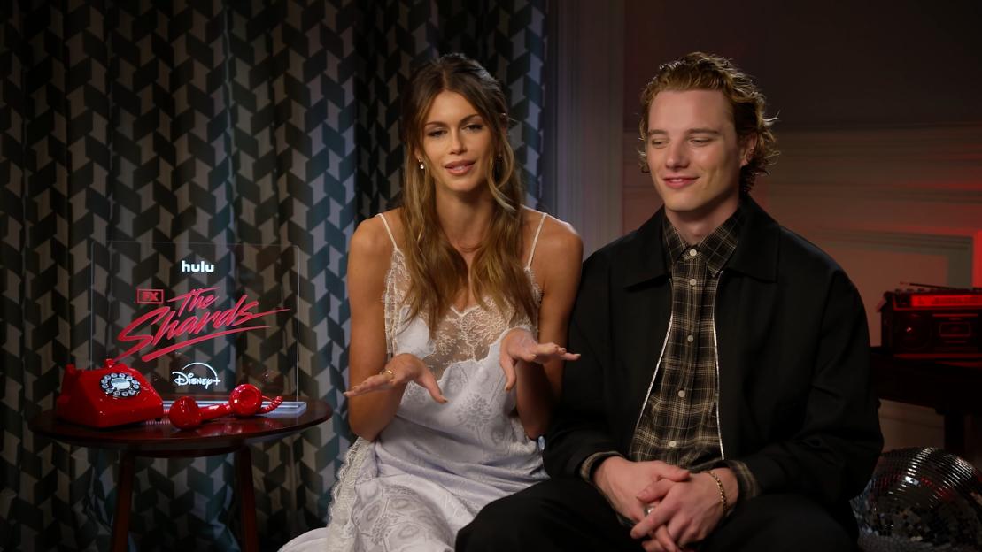 Kaia Gerber and another individual sitting together, with a Hulu, FX, and Disney+ sign in the foreground.