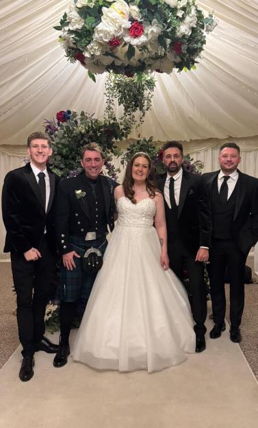 A bride in a white gown and four men in tuxedos, one in a kilt, stand before a floral arch.