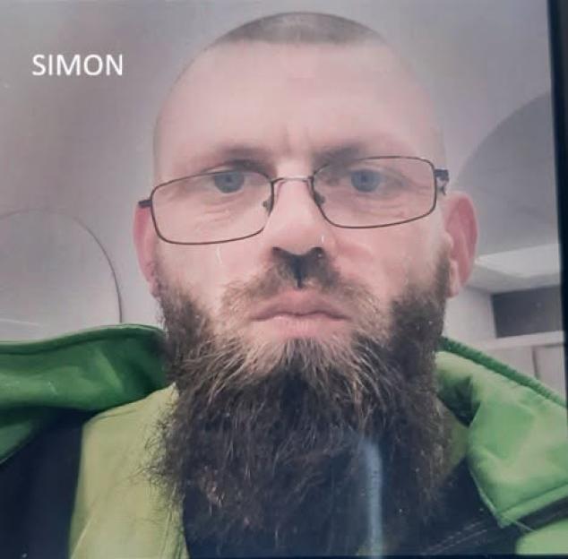 Close-up photo of a man named Simon with a bald head, glasses, and a full beard, wearing a green jacket.