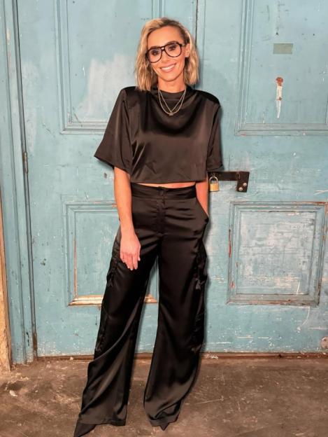 A woman with blonde hair and glasses poses in a black satin crop top and pants.