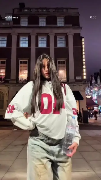 A young woman with long dark hair, wearing a white jersey with red lettering, light-wash jeans, and holding a water bottle, poses at night on a city street.