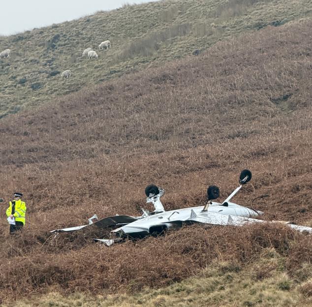 An emergency services worker in a high-visibility jacket stands next to a crashed light aircraft, which lies upside down on a brown, grassy hillside.