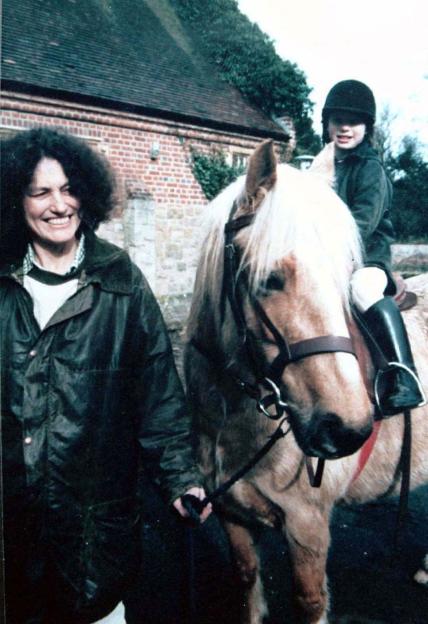 Lin Russell, smiling, leads a horse with her daughter Megan, wearing a riding helmet, sitting on its back.