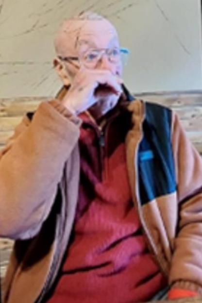 A bald man wearing glasses, a brown and black jacket, and a red shirt, with his hand near his mouth.