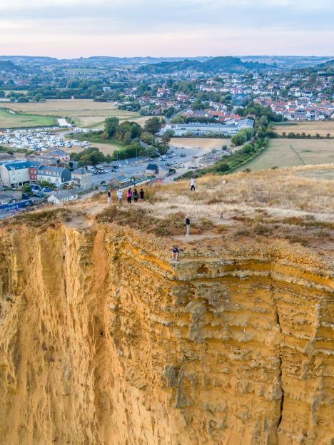 A teenage boy sits on the edge of a crumbling 150ft cliff overlooking a town with a crack visible in the rock face beneath him.