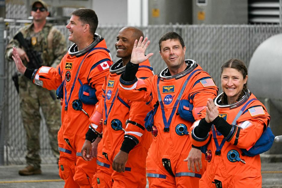 The Artemis II crew, consisting of three men and one woman in orange space suits, wave and smile as they walk.