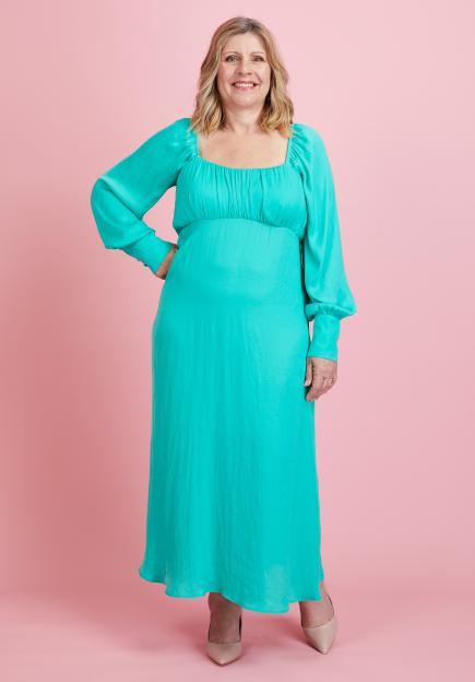 Kari Roberts, a woman with blonde hair, wearing a teal dress, smiles at the camera with her hand on her hip.