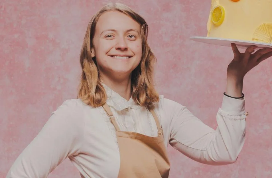 Danni, a contestant from The Great British Bake Off, smiling while holding up a yellow lemon cake against a pink background.