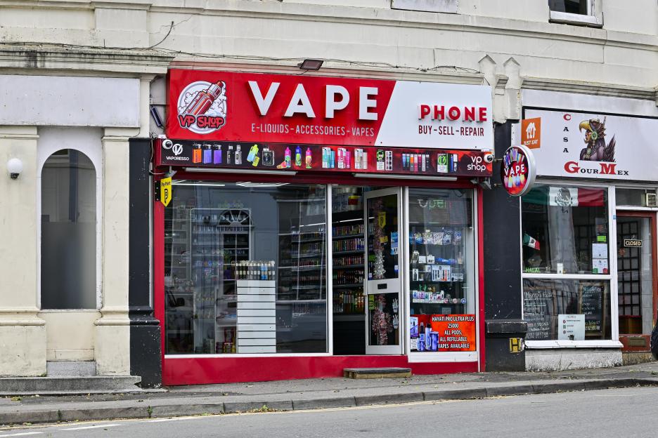 A vape shop with a large red and white sign for "VAPE E-LIQUIDS - ACCESSORIES - VAPES" and "PHONE BUY - SELL - REPAIR".