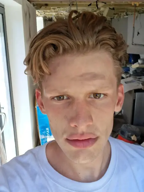 Close-up of a young man with brown hair and light stubble wearing a white t-shirt, looking directly at the camera.