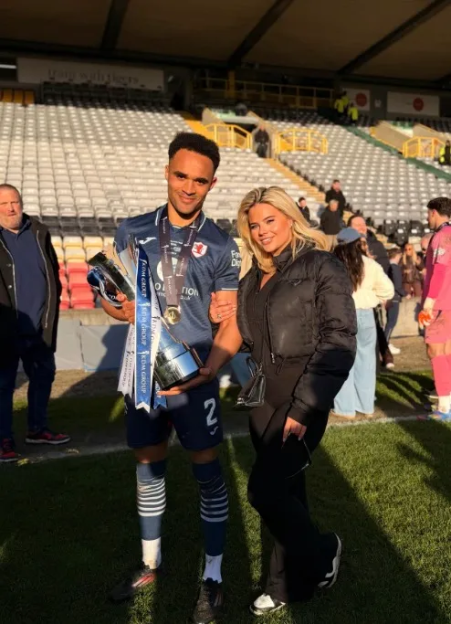 A male soccer player in uniform holds two trophies and stands next to a woman in a black jacket and pants on a soccer field.
