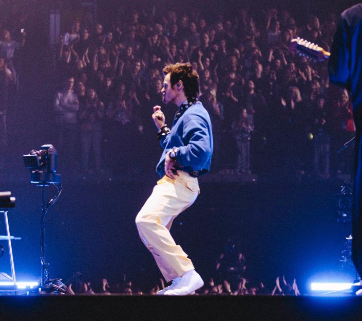 Harry Styles performing live on stage with his band, with an audience in the background.