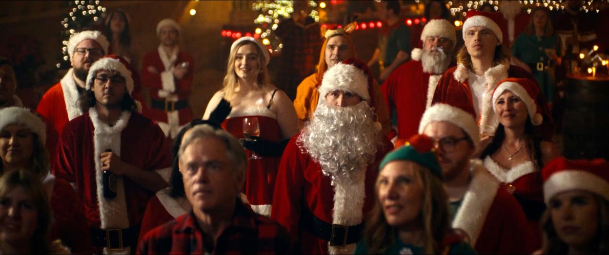 Rohan Campbell as Billy Chapman in a crowd of people dressed in Santa Claus costumes, some with drinks in hand.