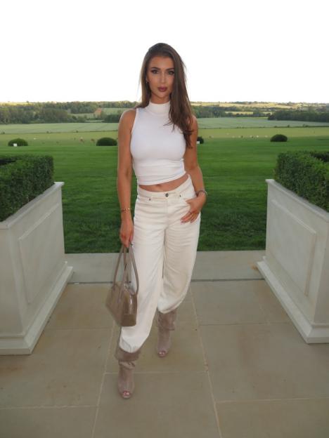 A woman in a white sleeveless top, cream pants, and open-toe boots stands on a patio with a large green field and distant trees behind her.