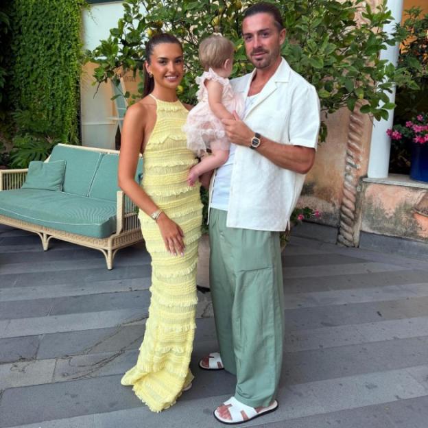 A woman in a yellow dress, a man in a white shirt and green pants, and a baby in a pink dress standing together.