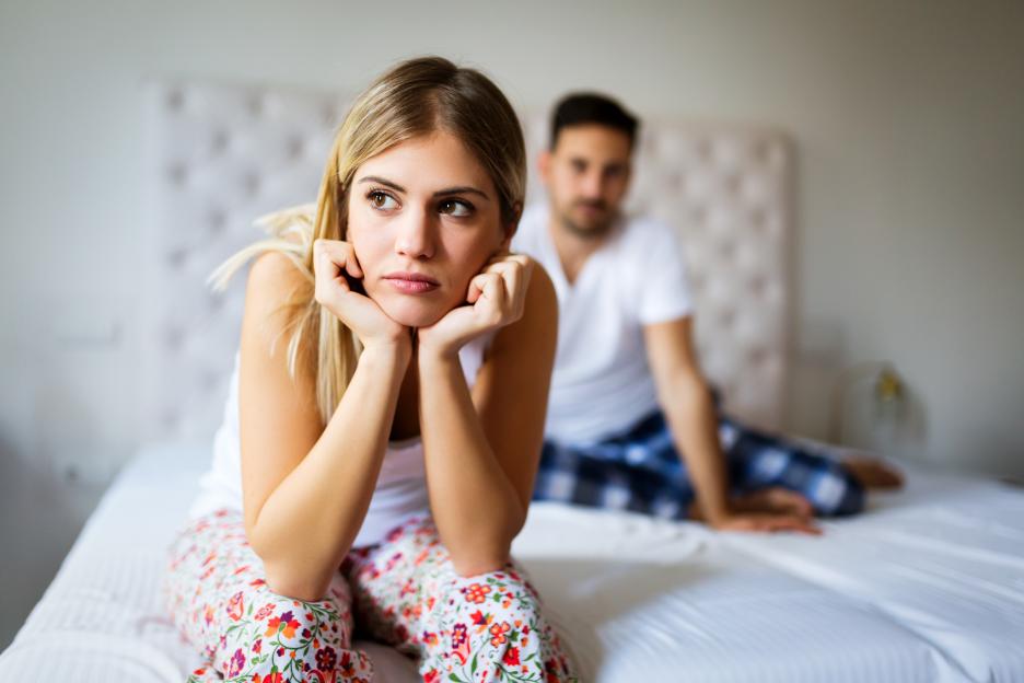 Unhappy woman sitting on bed with her head in her hands while man sits behind her.