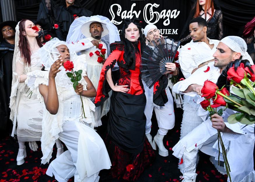 Lady Gaga in a red and black outfit with a black fan, surrounded by individuals in white outfits holding red roses.