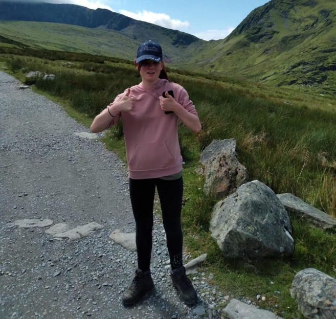 Holly Newton giving a thumbs-up while standing on a path in a mountainous landscape.