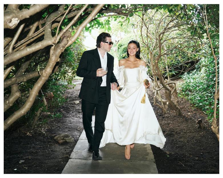Brooklyn Beckham and Nicola Peltz walking on a path surrounded by trees and greenery.