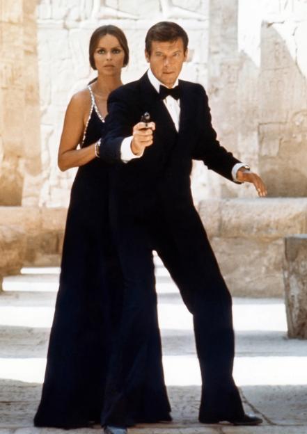 Roger Moore aiming a pistol while Barbara Bach stands behind him, both dressed in formal attire in front of ancient ruins.