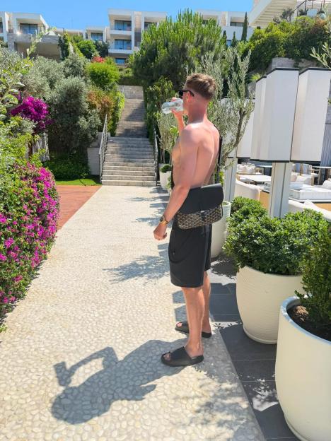 Cole Palmer drinks from a water bottle on a sunny path lined with flowers and greenery.