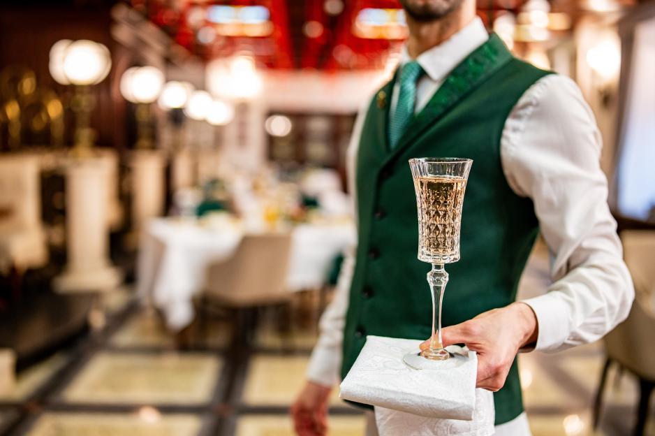 A waiter in a white shirt and green vest holds a glass of champagne with a white napkin.