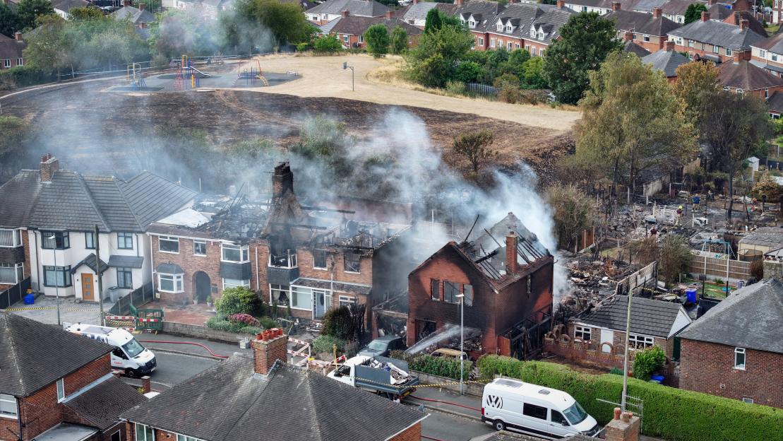 Aerial view of fire-damaged homes and a burnt landscape in Stoke on Trent, England, with smoke still rising from the ruins.