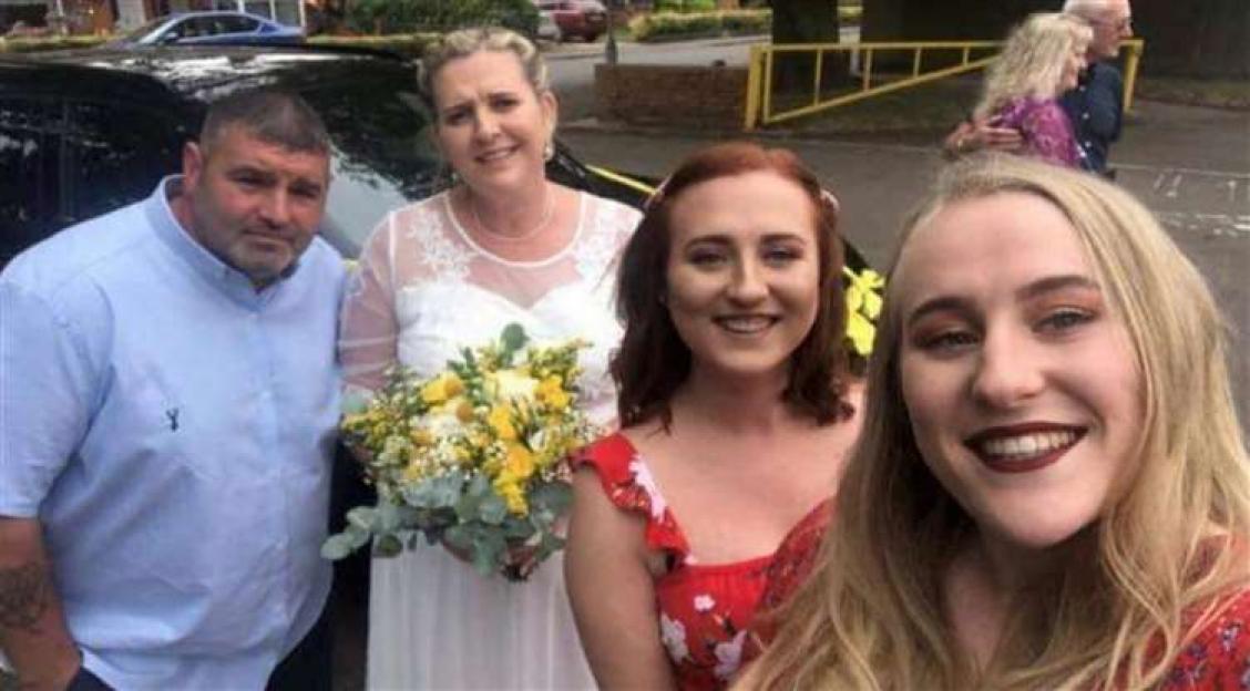 A man in a light blue shirt, a woman in a white wedding dress holding a bouquet, and two young women in red dresses smiling for a picture.