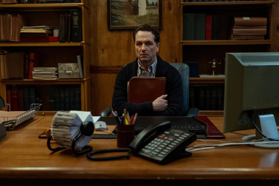 A man sits at a wooden desk in front of bookshelves, holding a brown folder.