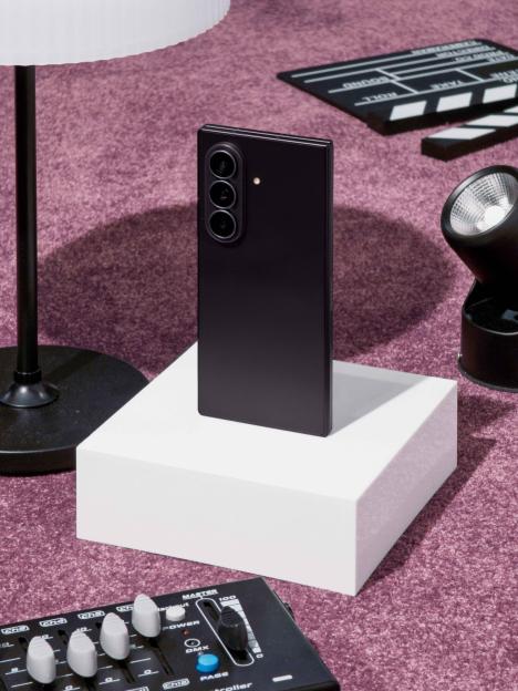 A black smartphone with a triple camera stands upright on a white block, surrounded by a clapperboard, stage light, audio mixer, and lamp, all on a purple carpet.