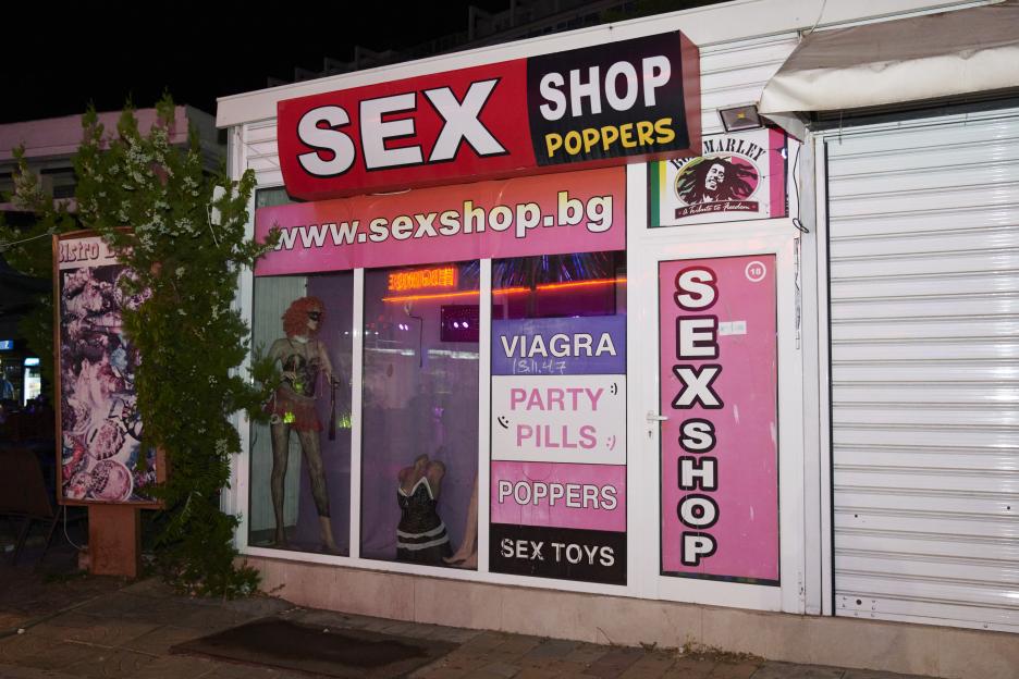 An exterior view of a sex shop with products like viagra, party pills, poppers, and sex toys advertised on the windows.