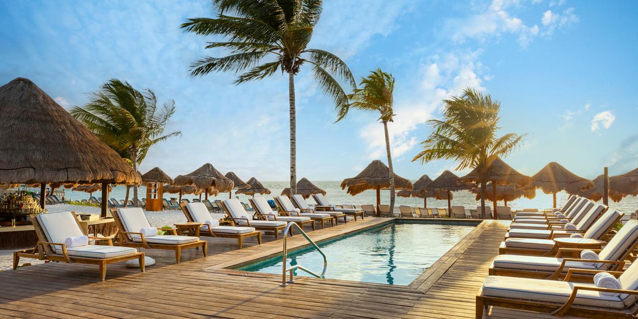 The Fairmont Mayakoba hotel in Riviera Maya with a poolside deck, lounge chairs, palm trees, and straw-roofed palapas on a beach by the ocean.