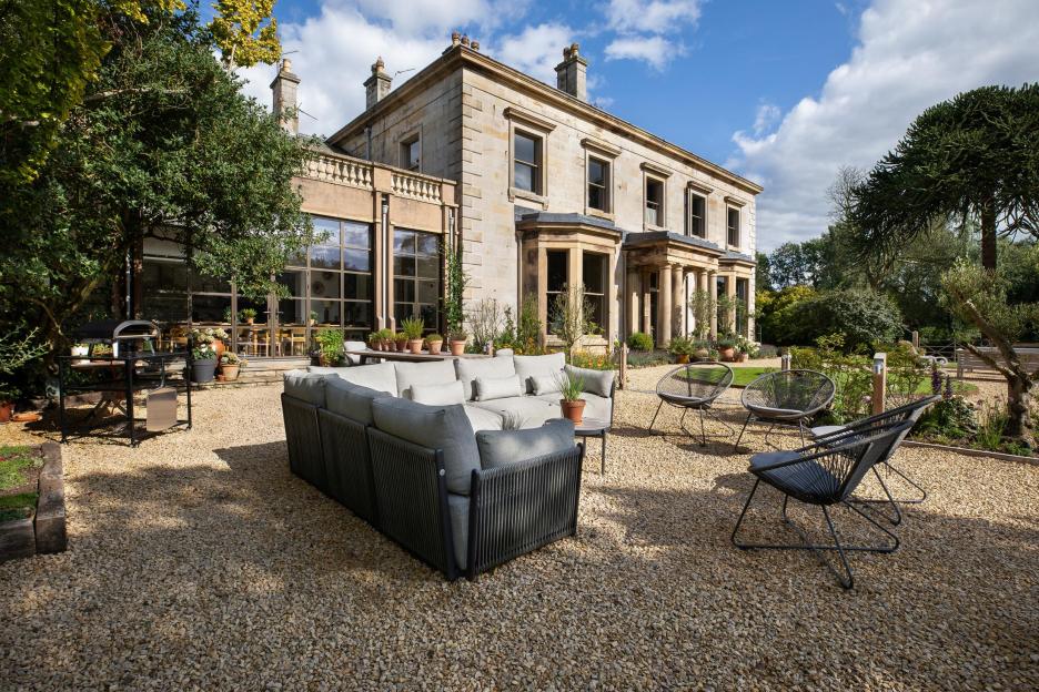 Outdoor patio with a large stone house in the background and a gray sectional sofa and chairs on a gravel ground in the foreground.