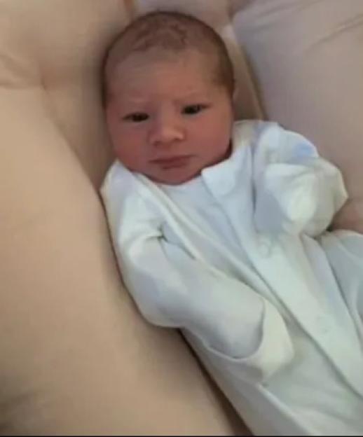 A newborn baby in a white onesie lies on a light pink cushion.