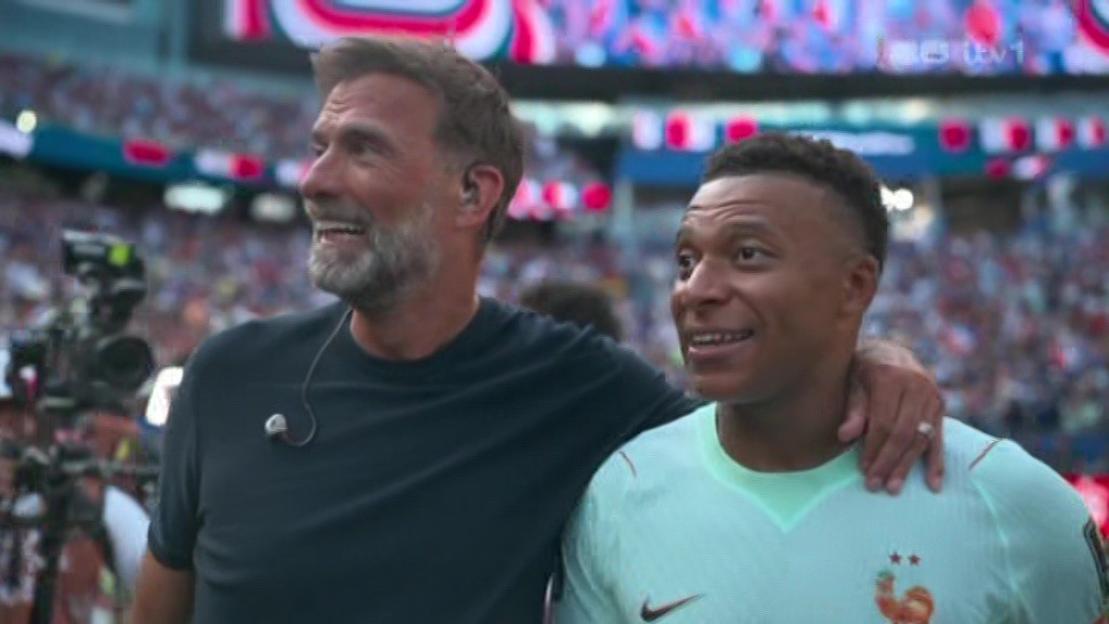 Two men smiling in a crowded stadium, one with his arm around the other.