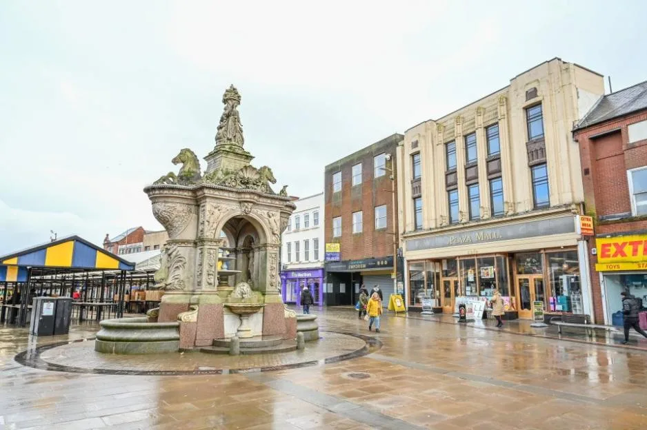 A stone fountain in a nearly empty town square in Dudley, with a few people walking past closed shops and the Plaza Mall.