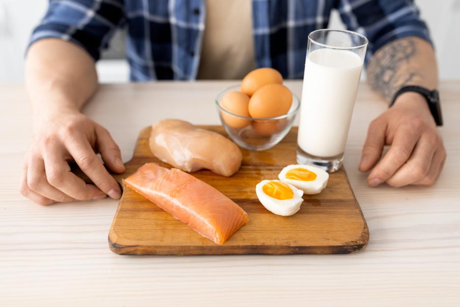 A person's hands rest on a table next to a wooden board with raw chicken breast, salmon fillet, and two halved boiled eggs. A bowl of whole eggs and a glass of milk are also on the table.