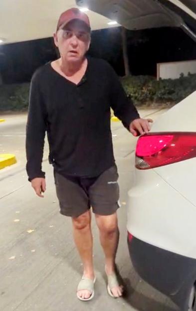 Shane Richie, wearing a black shirt, shorts, and sandals, standing next to a white car at a petrol station at night.