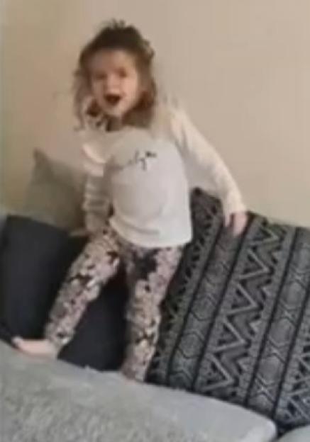 A young girl in a white shirt and patterned pants stands on a couch cushion.
