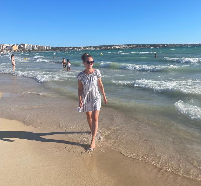 A woman in a striped dress walks barefoot on a sandy beach as waves crash around her.