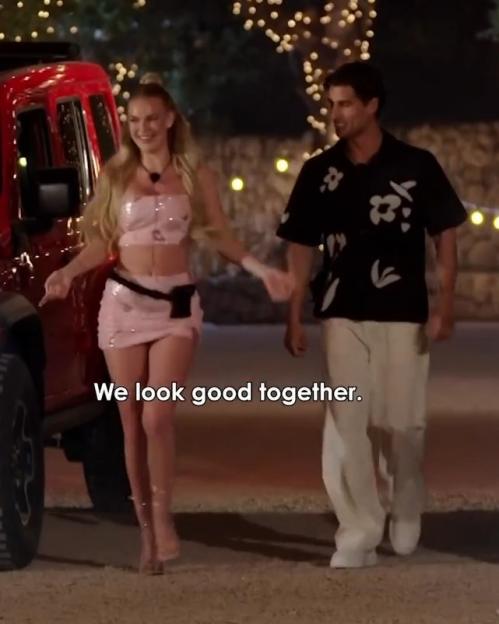 A woman in a pink outfit and a man in a black and white shirt walk hand-in-hand next to a red car at night.