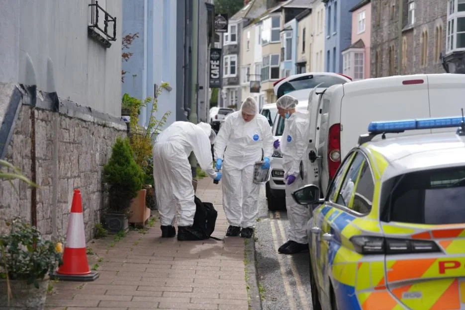 Forensic investigators in white protective suits working on a street alongside police vehicles and emergency vans.